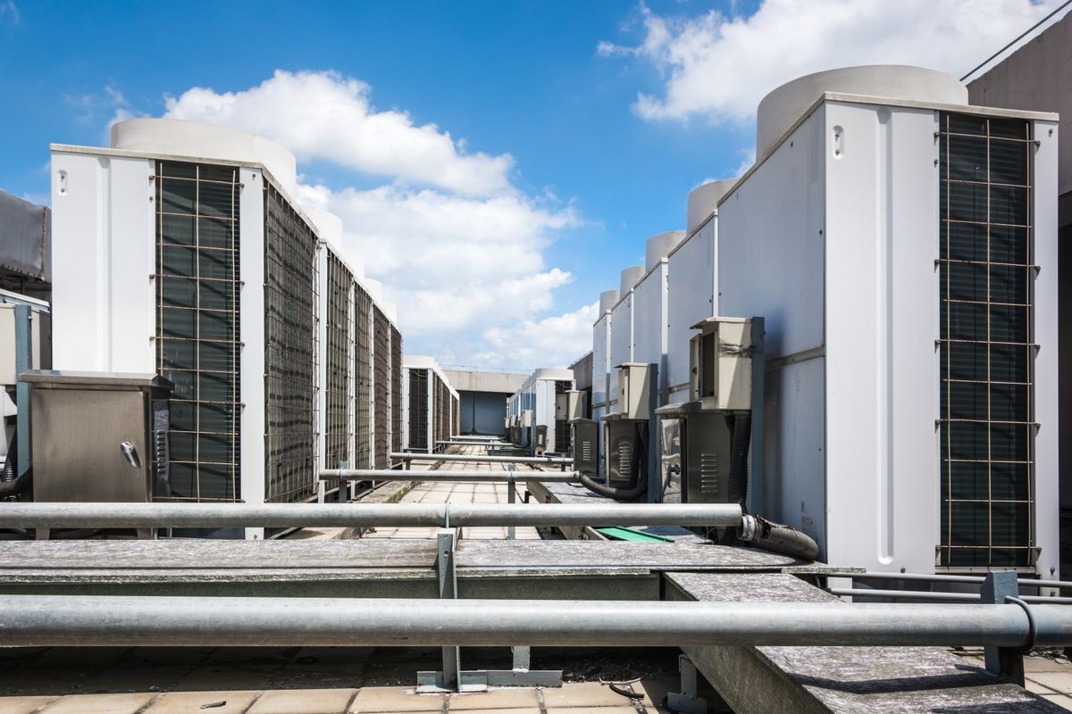 Square air-conditioning unit on the roof with a round fan. In the background gradually receding other units that are out of focus. On the right side light blue sky and commercial space.